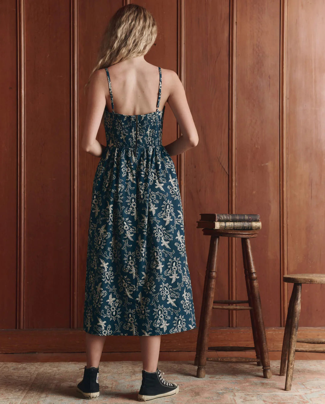 A woman with long blond hair faces a wooden wall, wearing The Great Inc.'s blue and white cotton Verse Dress with a sculpted silhouette and black sneakers. Beside her are a tall stool stacked with books and a shorter wooden bench.