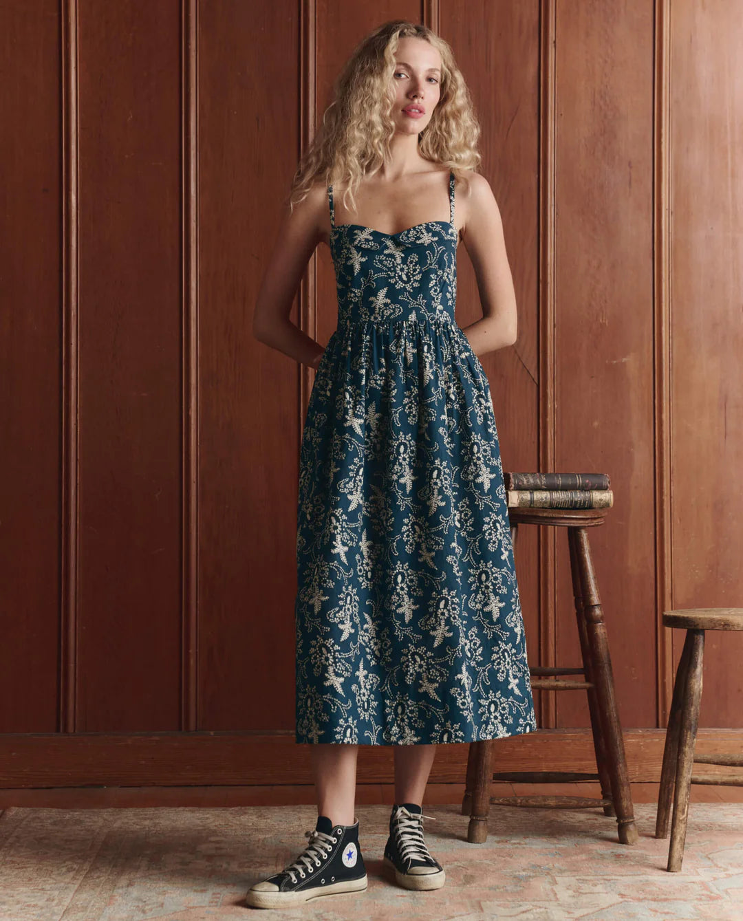 A woman with long blonde hair stands indoors against wood paneling, wearing The Verse Dress by The Great Inc.—a blue cotton dress with a floral pattern and sculpted silhouette—paired with black high-top sneakers. She rests a hand on books atop a wooden stool.