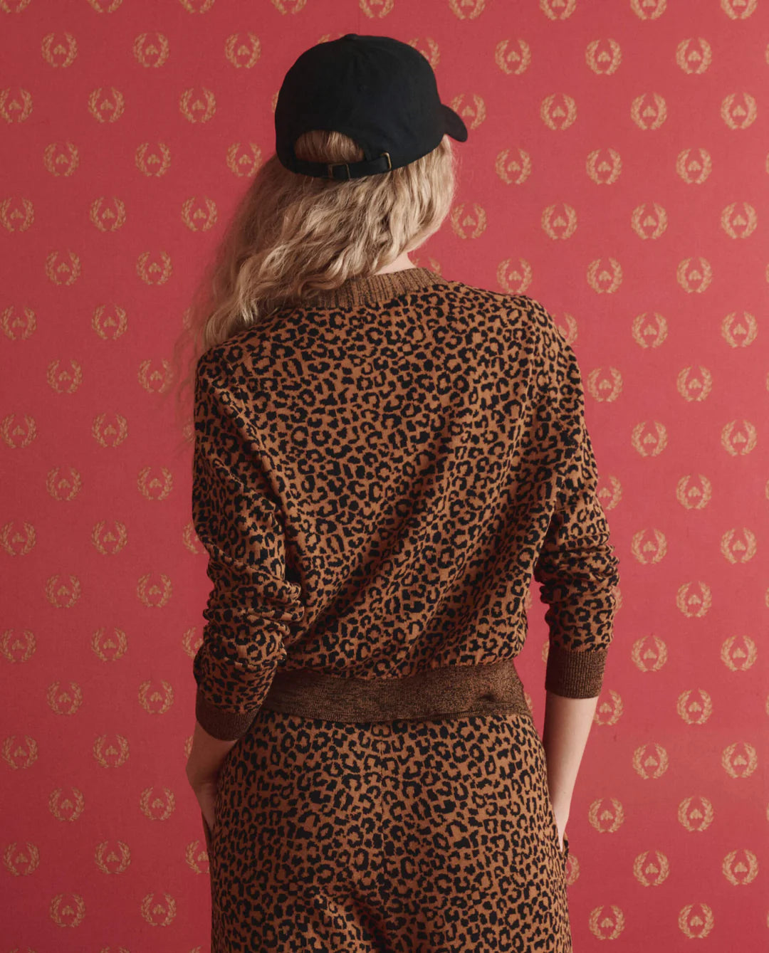 A person with long, wavy blonde hair, wearing a black cap and The Great Inc. Shrunken Vneck Cardigan, stands facing a red patterned wall, shown from behind.