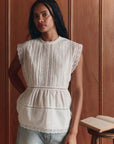 A woman with long dark hair wears The Sonnet Top by The Great Inc.—a sleeveless Victorian-inspired blouse with delicate lace—paired with light blue jeans, standing before a wooden paneled wall beside an open book.