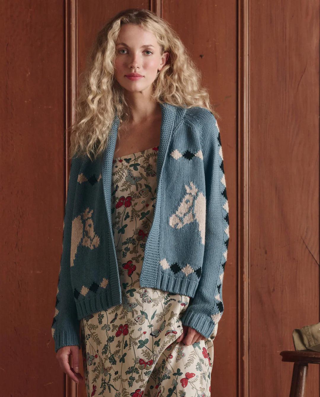 A woman with long curly blonde hair wears The Great Inc. Stable Lodge Cardigan, featuring a white western horse motif over a floral dress, standing before a wooden-paneled wall.