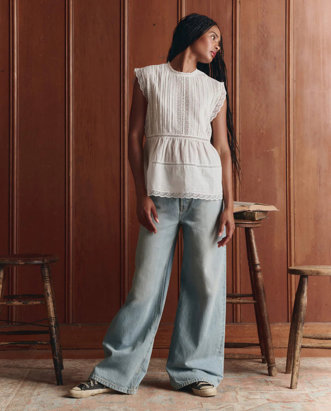 A person with long braided hair wears The Sonnet Top by The Great Inc.—a Victorian-inspired piece with delicate lace—paired with wide-leg light blue jeans, standing indoors between two wooden stools against a paneled wooden wall.