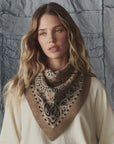 A woman with long wavy blonde hair wears The Bandana Kerchief by The Great Inc., featuring a brown alpaca knit with a black and white paisley pattern, along with a cream sweater, posed before a textured gray fabric backdrop.