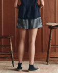 Indoors by a wooden wall, a person wears The Boxer Short by The Great Inc.—black and white vertical striped pull-on sleep shorts—paired with a navy top and black high-top sneakers, flanked by two wooden stools, hands on hips.