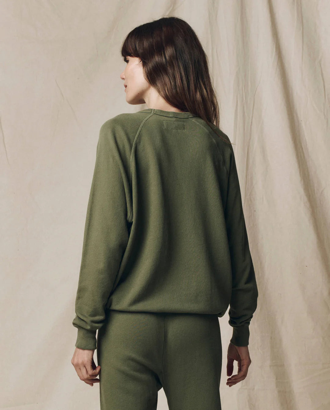 A woman with long brown hair wears The Great Inc.'s oversized College Sweatshirt S25 and matching pants, standing with her back to the camera against a beige fabric backdrop.
