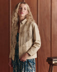 A woman with long curly blonde hair wears The Leather Fringe Jacket by The Great Inc. over a blue patterned dress, standing beside a stack of books against a wooden-paneled wall.