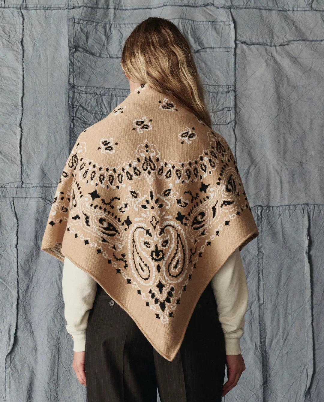 A person with long, wavy hair wears The Great Inc.'s The Big Bandana Scarf—beige with a black and white intricate pattern—draped over their shoulders, set against a quilted gray background.
