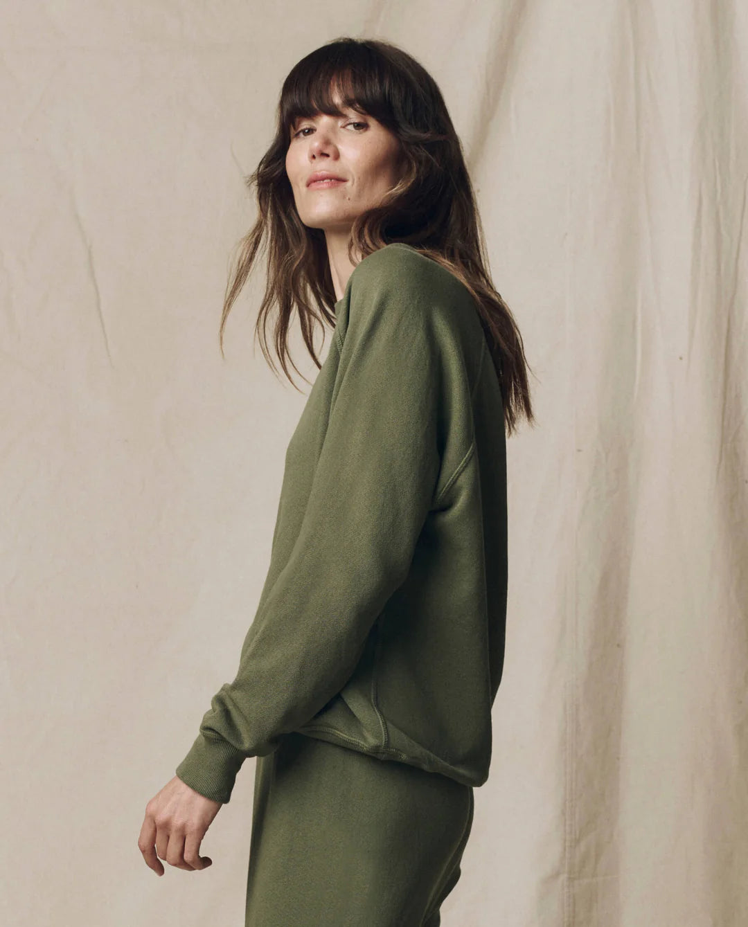 A person with long brown hair models The College Sweatshirt S25 by The Great Inc. in olive green, styled with matching pants, standing sideways and looking at the camera against a neutral beige fabric background.