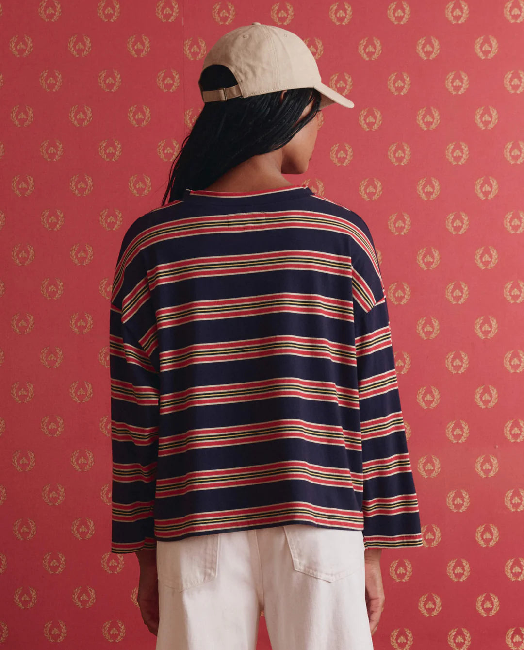 A person stands by a red patterned wall wearing The Great Inc.'s The Field Tee—a handcrafted navy long-sleeve with red and white stripes—paired with a beige cap and white pants. Their hair is visible beneath the cap.