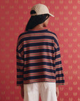 A person stands by a red patterned wall wearing The Great Inc.'s The Field Tee—a handcrafted navy long-sleeve with red and white stripes—paired with a beige cap and white pants. Their hair is visible beneath the cap.