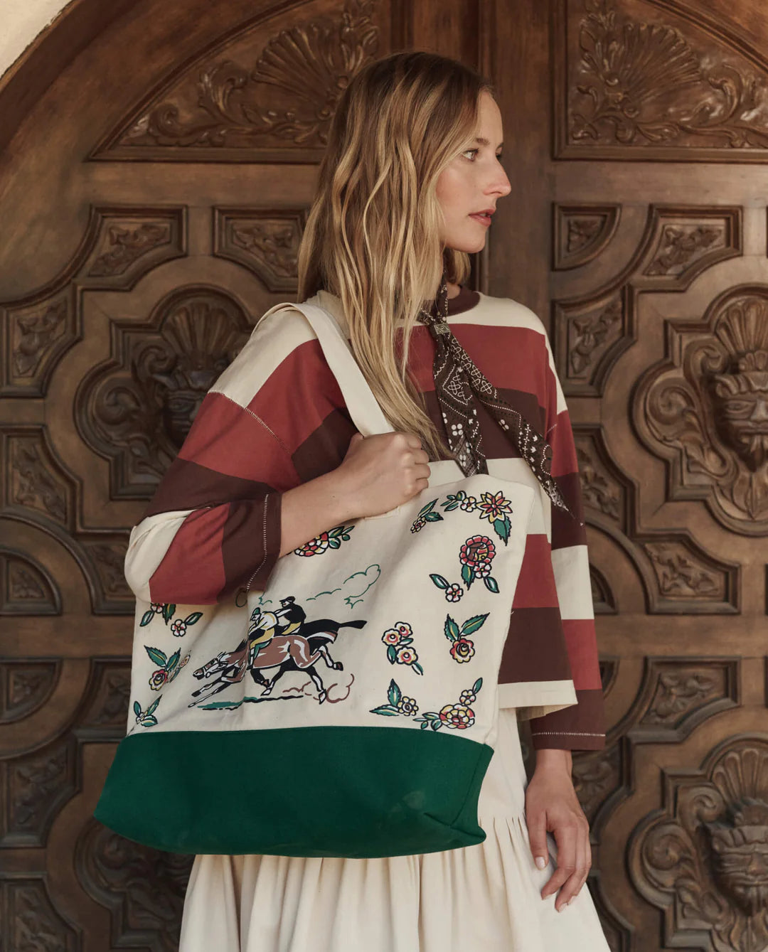 A woman with wavy blonde hair stands before an ornate wooden door, wearing a striped top and cream skirt. She carries The Derby Tote by The Great Inc., featuring a green base and colorful embroidered design.