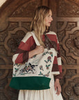 A woman with wavy blonde hair stands before an ornate wooden door, wearing a striped top and cream skirt. She carries The Derby Tote by The Great Inc., featuring a green base and colorful embroidered design.