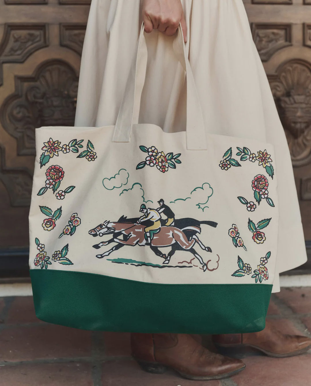 A person in a cream skirt holds The Derby Tote by The Great Inc., a cotton canvas bag with floral designs, horse-racing jockey illustration, and a dark green bottom, contrasting with an ornate wooden door and stone tiles.