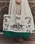 A person in a cream skirt holds The Derby Tote by The Great Inc., a cotton canvas bag with floral designs, horse-racing jockey illustration, and a dark green bottom, contrasting with an ornate wooden door and stone tiles.