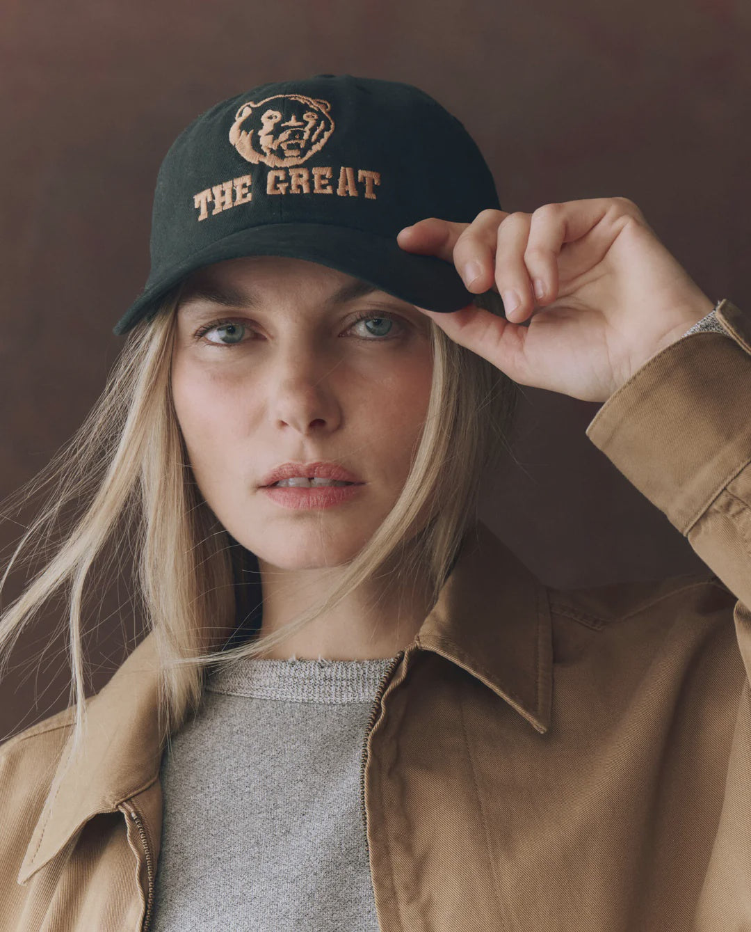A person with long blonde hair wears The Great Inc.'s The Baseball Hat—a black cap with vintage chainstitch "THE GREAT" and a lion graphic—paired with a tan jacket and gray top, looking at the camera while holding the brim.