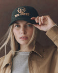 A person with long blonde hair wears The Great Inc.'s The Baseball Hat—a black cap with vintage chainstitch "THE GREAT" and a lion graphic—paired with a tan jacket and gray top, looking at the camera while holding the brim.