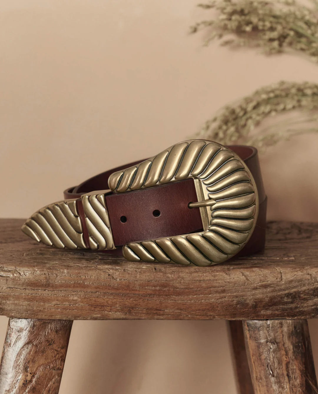 The Big Buckle Belt by The Great Inc., made from top-grain leather with a large fluted buckle featuring textured leaf-like patterns, is coiled on a rustic wooden stool against a beige background with dried grasses.