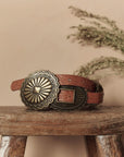 The Great Inc. Leather Concho Belt, featuring engraved patterns and a large ornate buckle, is displayed on a rustic wooden stool with blurred foliage in the background, highlighting its authentic Western craftsmanship.