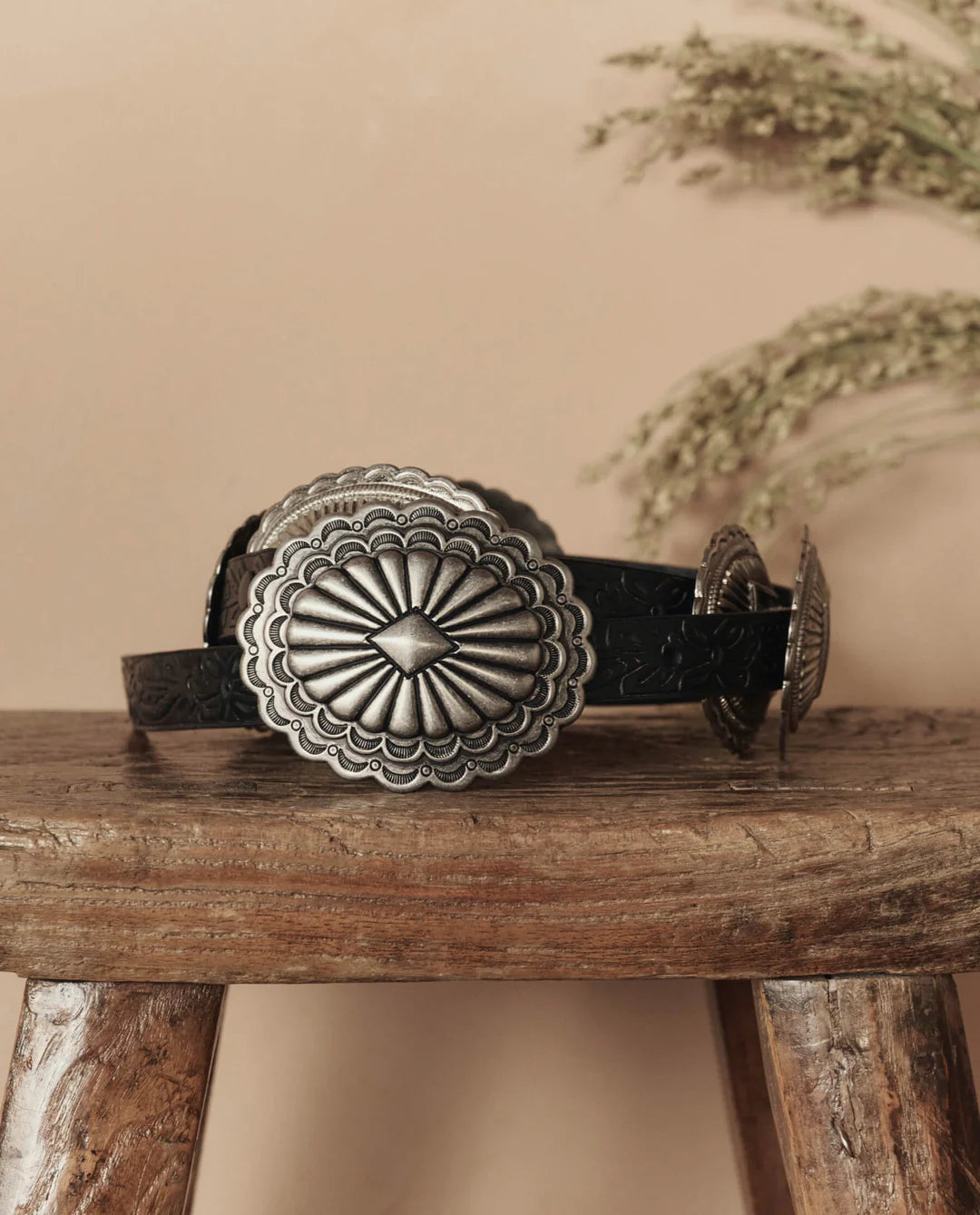 The Great Inc. Leather Concho Belt, crafted from top-grain leather with large, ornate silver conchos, sits on a rustic wooden stool with dried greenery and a beige wall in the background—showcasing true Western craftsmanship.