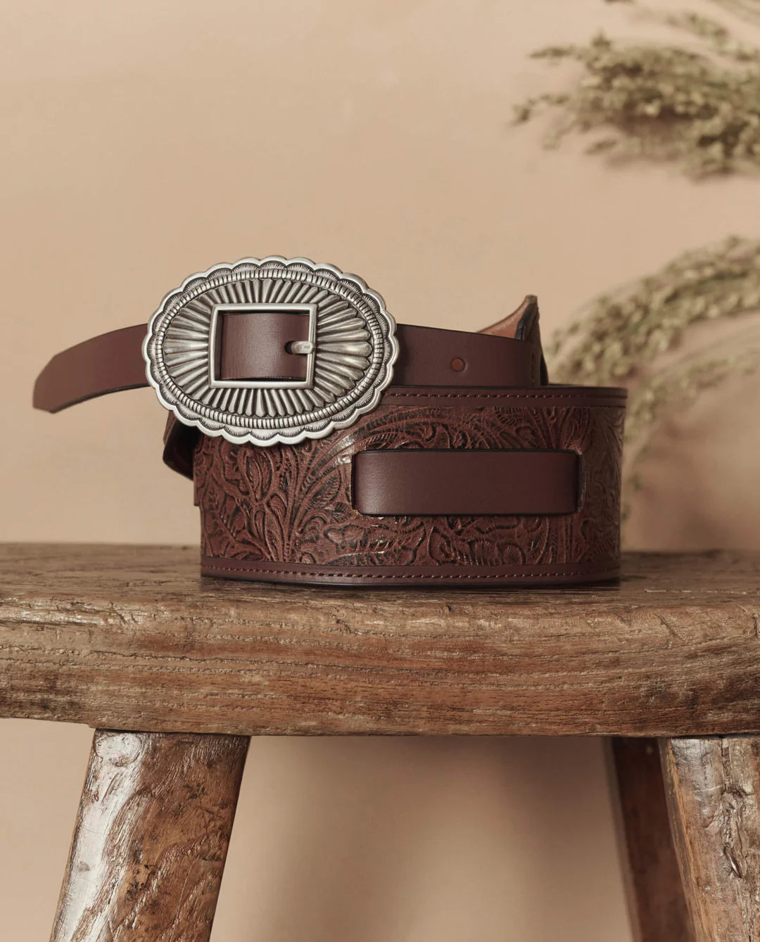 The Great Inc. Tooled Waist Belt, crafted from top-grain leather with an ornate silver buckle and embossed floral patterns, rests coiled on a rustic wooden stool with dried foliage in the background.