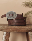 The Great Inc. Tooled Waist Belt, crafted from top-grain leather with an ornate silver buckle and embossed floral patterns, rests coiled on a rustic wooden stool with dried foliage in the background.
