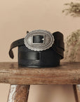 The Great Inc. Tooled Waist Belt with an ornate silver buckle rests on a rustic wooden stool, set against a beige background with blurred dried flowers in the corner.