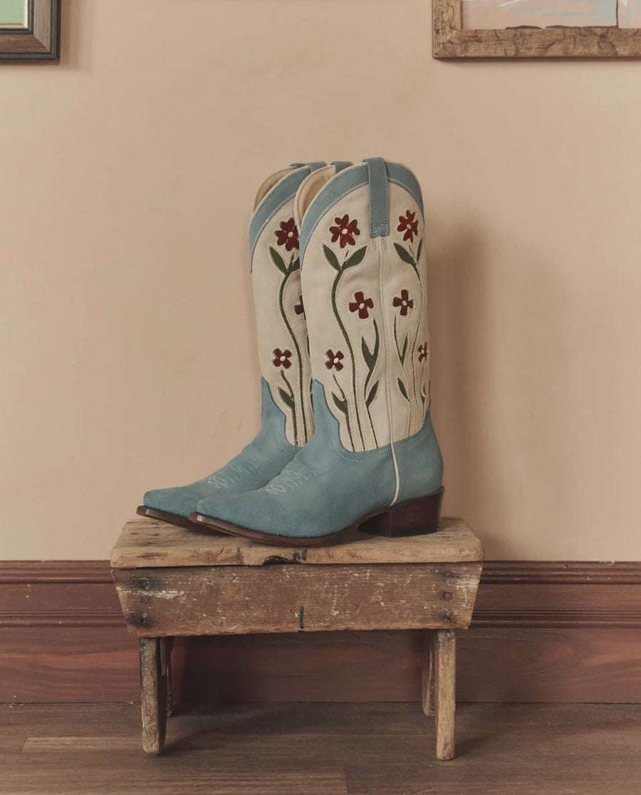 The Great Inc. Suede Floral Cowboy Boot featuring red and green embroidery is displayed on a rustic wooden stool against a beige wall.