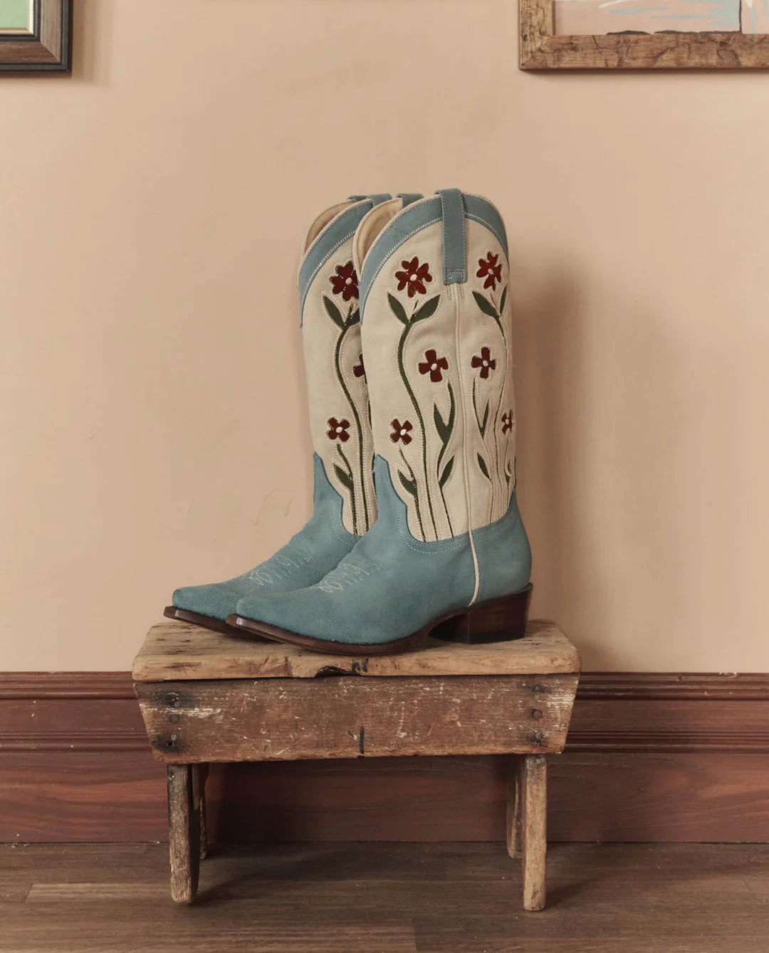 The Great Inc. Suede Floral Cowboy Boot, in vintage blue with cream and red floral embroidery, is displayed on a rustic wooden stool against a beige wall with wood trim and framed pictures in view.