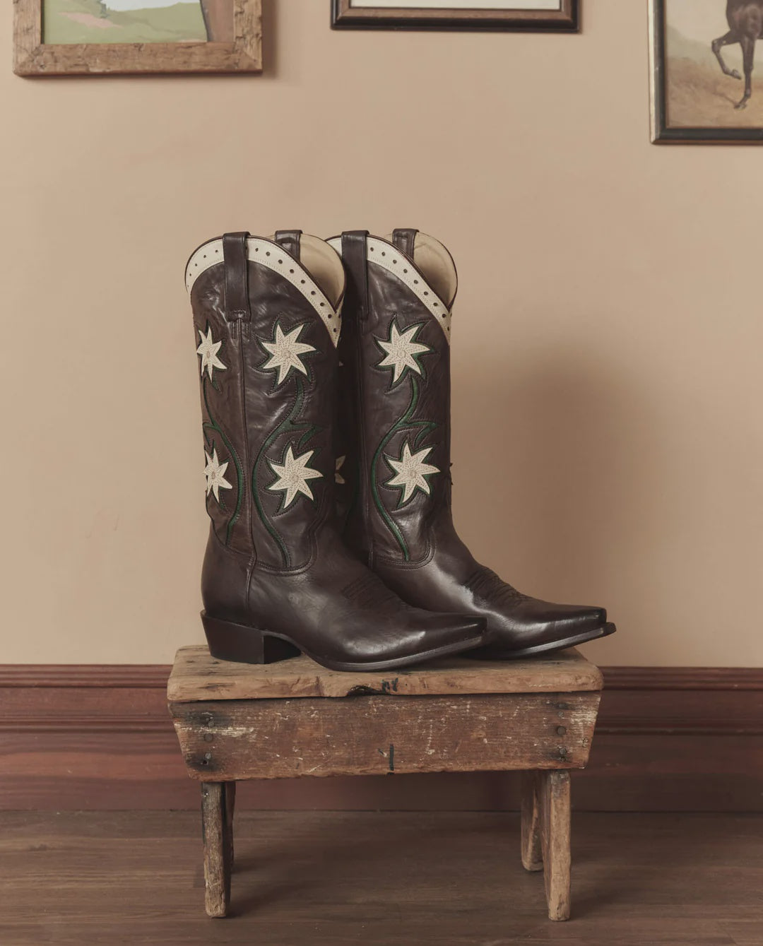 The Leather Floral Cowboy Boot by The Great Inc. features handcrafted leather with white star designs and pointed toes, displayed on a rustic wooden stool to add classic western style against a beige wall with framed pictures.