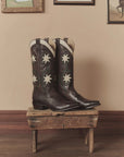 The Leather Floral Cowboy Boot by The Great Inc. features handcrafted leather with white star designs and pointed toes, displayed on a rustic wooden stool to add classic western style against a beige wall with framed pictures.