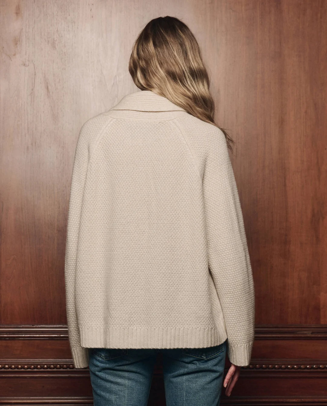 Cashmere cable-knit lodge cardigan by The Great Inc. in cream, styled with blue jeans against a rustic wood wall.
