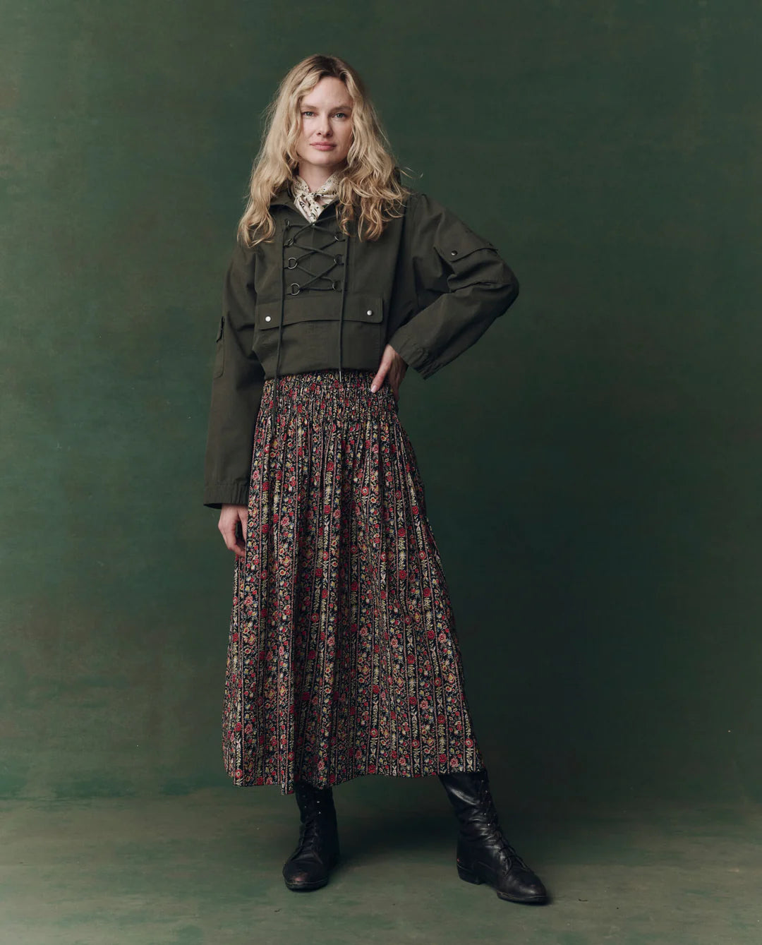 A woman with long blonde hair poses against a green background, wearing The Great Inc.’s The Canary Skirt with floral pleats, an olive cropped jacket, white blouse, and black lace-up boots, her hand resting on her hip.