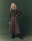 A woman with long blonde hair poses against a green background, wearing The Great Inc.’s The Canary Skirt with floral pleats, an olive cropped jacket, white blouse, and black lace-up boots, her hand resting on her hip.