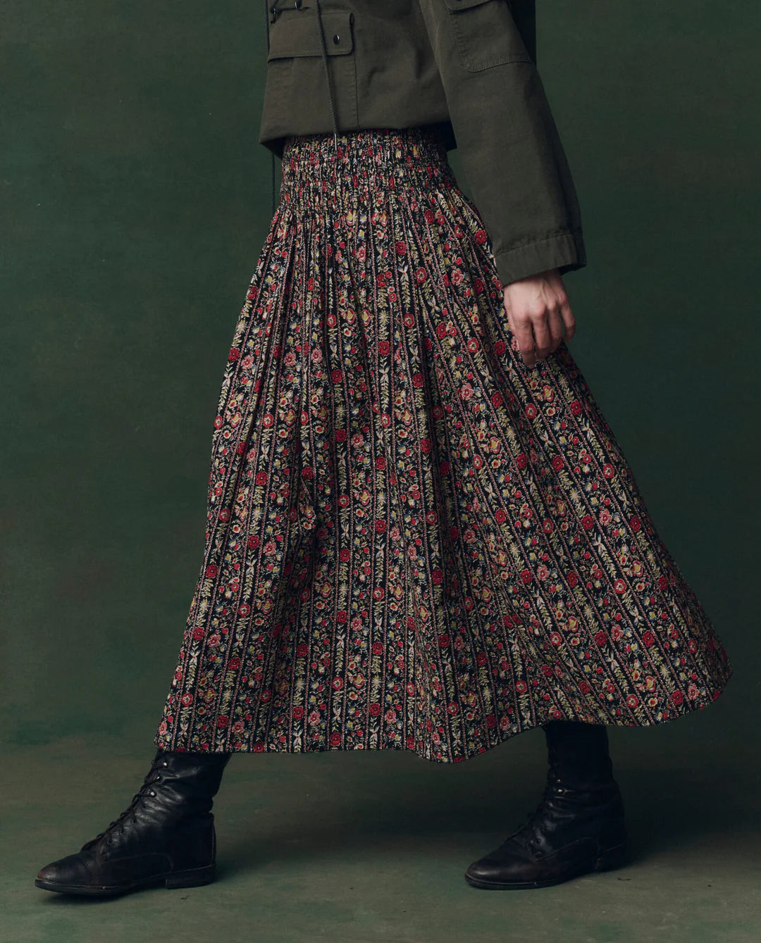 A person wearing The Canary Skirt by The Great Inc.—a long, pleated cotton seersucker skirt with a colorful floral pattern—plus a dark green jacket and black lace-up boots walks against a green background.