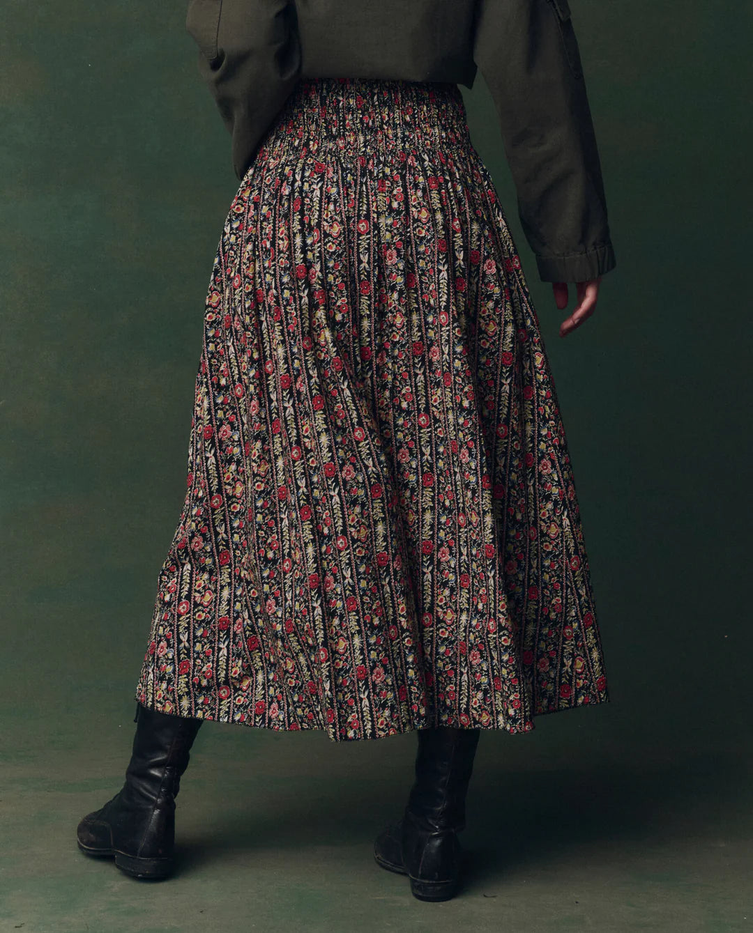 Someone wears The Canary Skirt by The Great Inc., an ankle-length cotton seersucker skirt with red and beige floral designs, paired with black boots and a dark green jacket, standing against a matching dark green background.