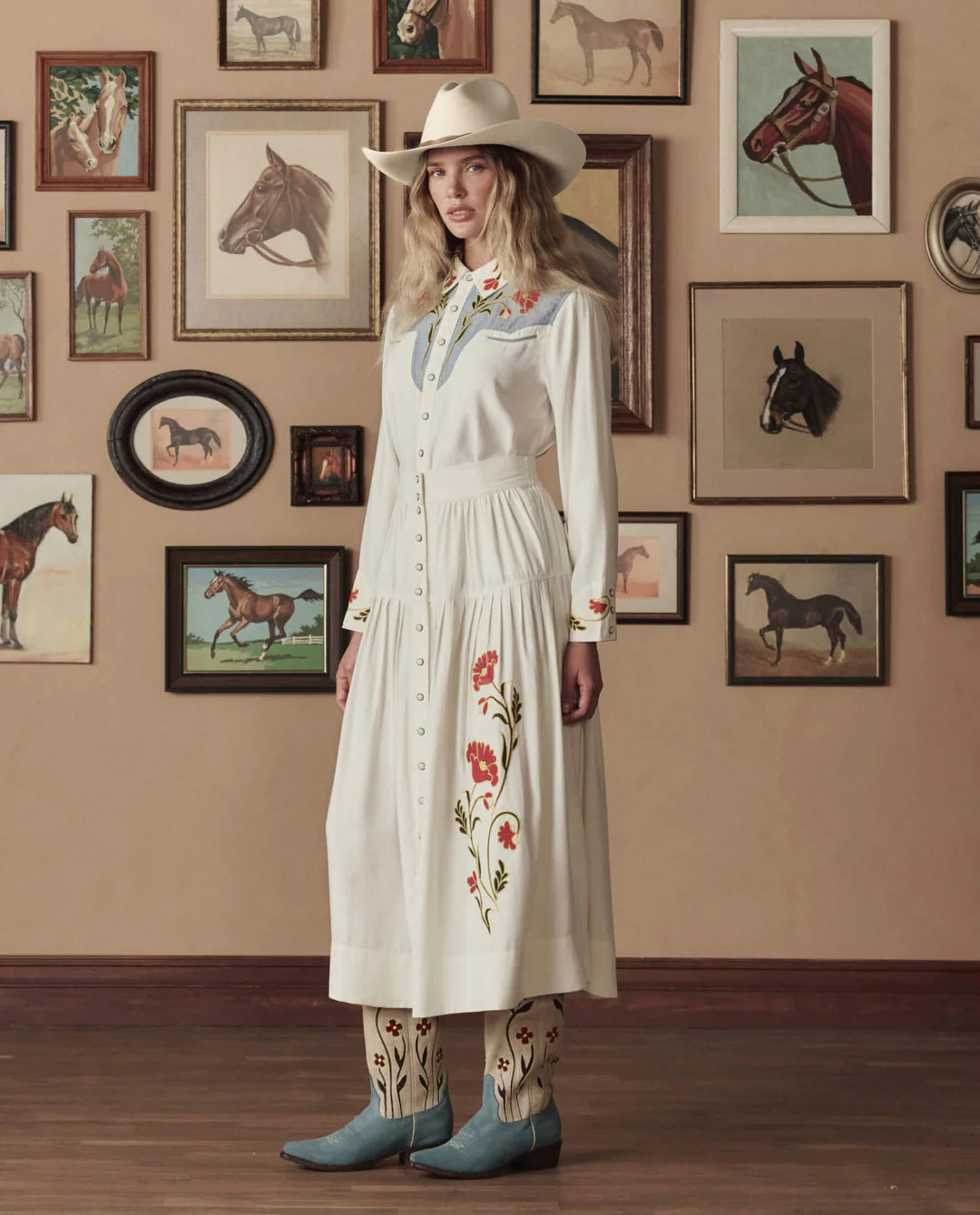 A woman in a white embroidered western dress, cowboy hat, and The Great Inc. Suede Floral Cowboy Boot stands indoors against a wall decorated with framed horse paintings and photos.