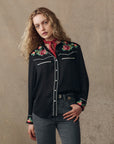 A person with wavy blonde hair wears The Great Inc.’s The Heirloom Rose Western Shirt—black with rose embroidery and pearl snaps—paired with a red neck scarf and gray jeans, standing against a plain beige background.