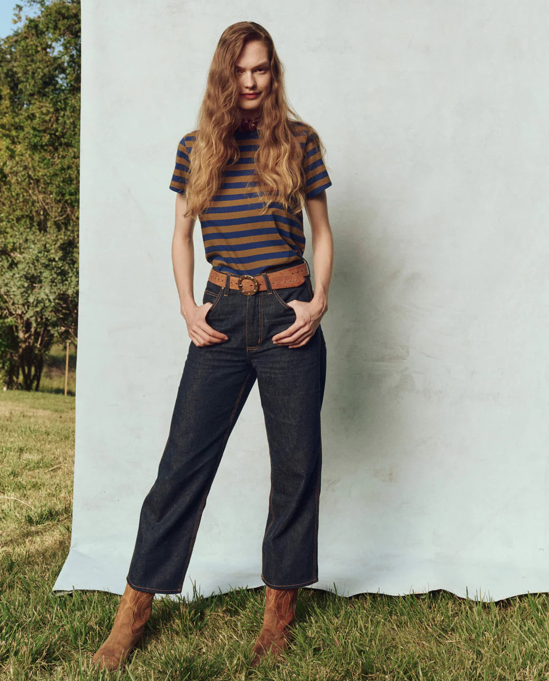 A person with long hair stands confidently outside against a light backdrop, wearing The Little Tee by The Great Inc., made from 100% cotton. Dark jeans, a brown belt, and brown boots complete the look amid lush grass and trees.