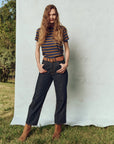 A person with long hair stands confidently outside against a light backdrop, wearing The Little Tee by The Great Inc., made from 100% cotton. Dark jeans, a brown belt, and brown boots complete the look amid lush grass and trees.