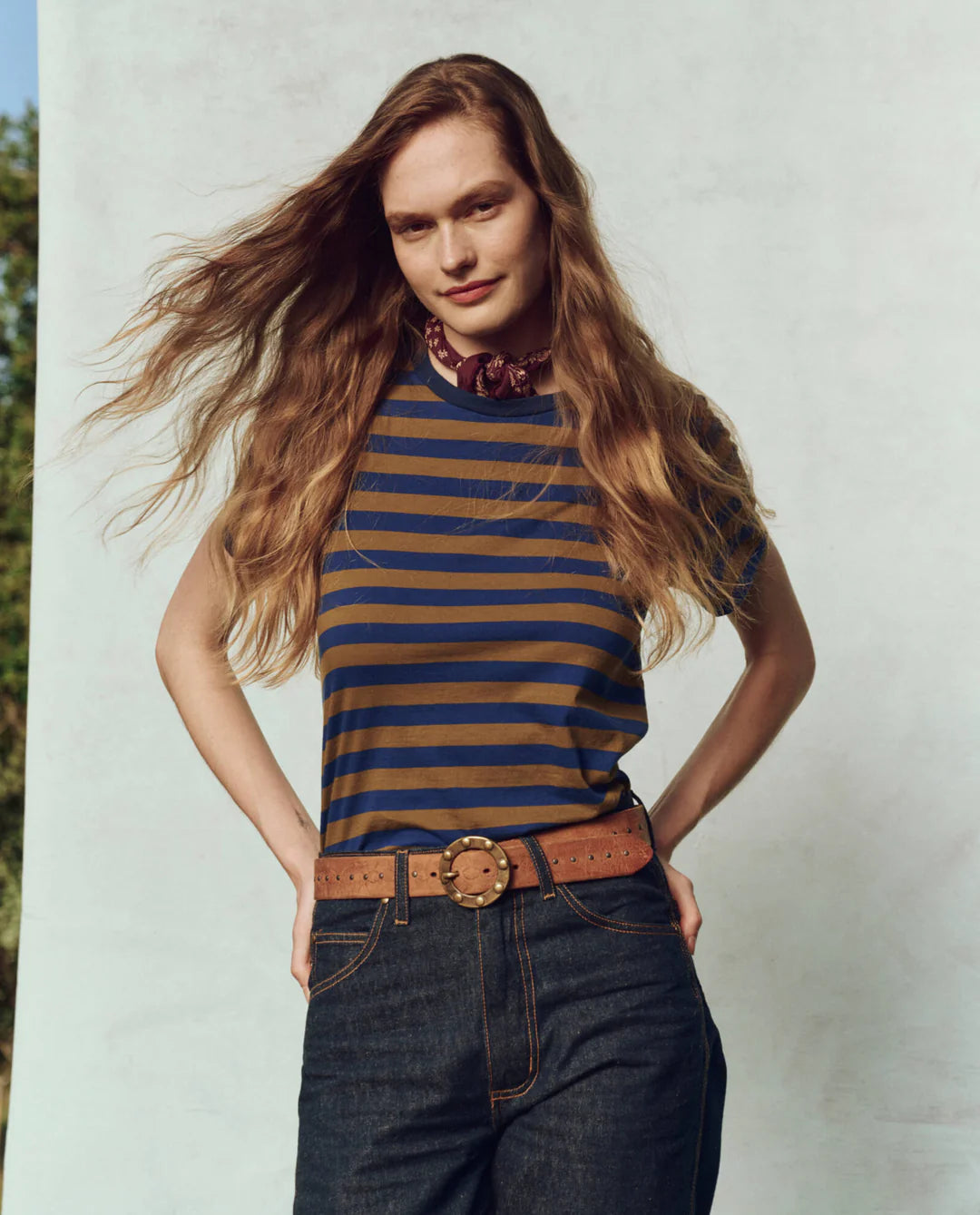 A person with long, wavy hair poses outdoors in The Little Tee by The Great Inc., paired with dark jeans and a brown belt. They stand against a light-colored background, with hands in their pockets and a slight smile on their face.