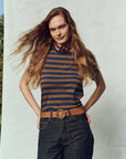 A person with long, wavy hair poses outdoors in The Little Tee by The Great Inc., paired with dark jeans and a brown belt. They stand against a light-colored background, with hands in their pockets and a slight smile on their face.