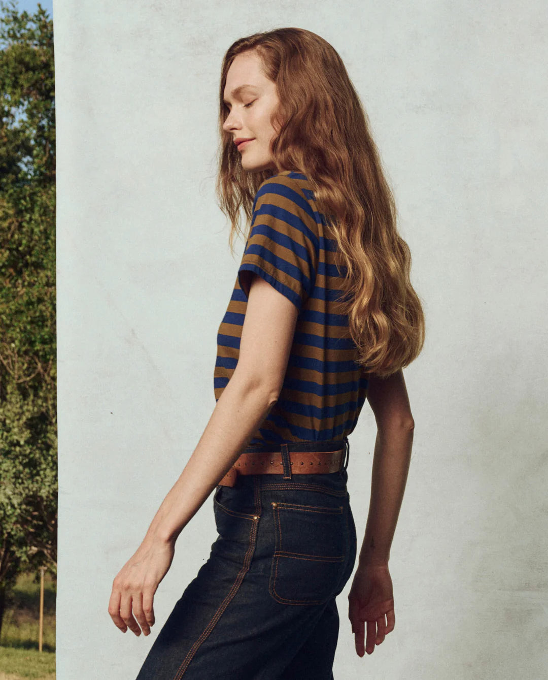 Against a light wall, a person with long, wavy hair stands sideways outdoors in jeans and The Little Tee by The Great Inc., with greenery visible in the background.