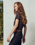 Against a light wall, a person with long, wavy hair stands sideways outdoors in jeans and The Little Tee by The Great Inc., with greenery visible in the background.