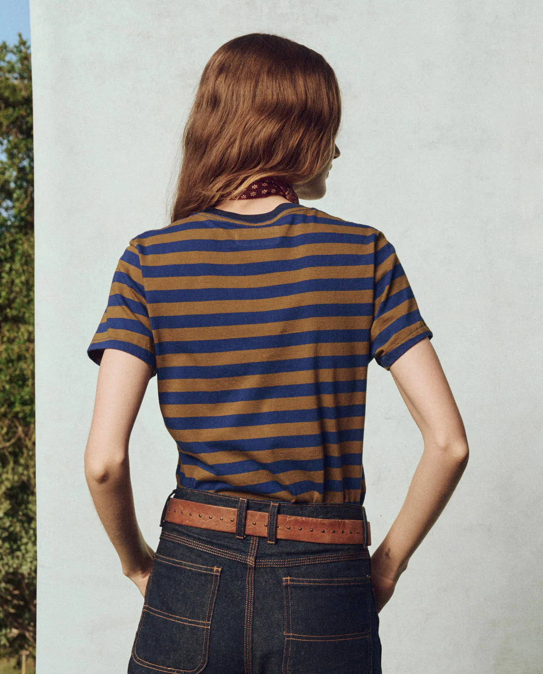 A person with long brown hair stands facing a wall, wearing The Little Tee by The Great Inc. and dark denim jeans. A brown belt and scarf around their neck complete the outfit. The 100% cotton fabric adds comfort and style, while green foliage is partially visible on the side.