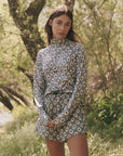 In a sunlit grove, a woman stands in The Quarter Zip Trail Pullover from The Great Inc., made of recycled polyester with white daisies on blue. The moisture-wicking fabric ensures comfort as she gazes at the camera, framed by leafy trees and grass.