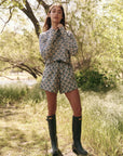 A person stands outdoors in a grassy area with trees, wearing a floral long-sleeved top and The Rover Short by The Great Inc. Their long hair flows over tall green boots as sunlight filters through the foliage.