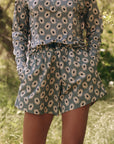 A person outdoors showcases outdoor clothing in a floral ensemble. The blue top and The Rover Short by The Great Inc. feature water-repellant shorts with white daisy patterns, perfectly complementing the lush, green wooded area in the background.