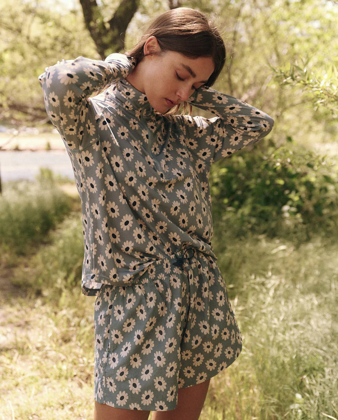 In a tranquil, green setting with lush foliage and trees, someone adjusts their hair while wearing The Great Inc.&#39;s daisy-patterned Rover Short, alongside a matching long-sleeve top. The shorts are designed to repel water, making them both stylish and functional.