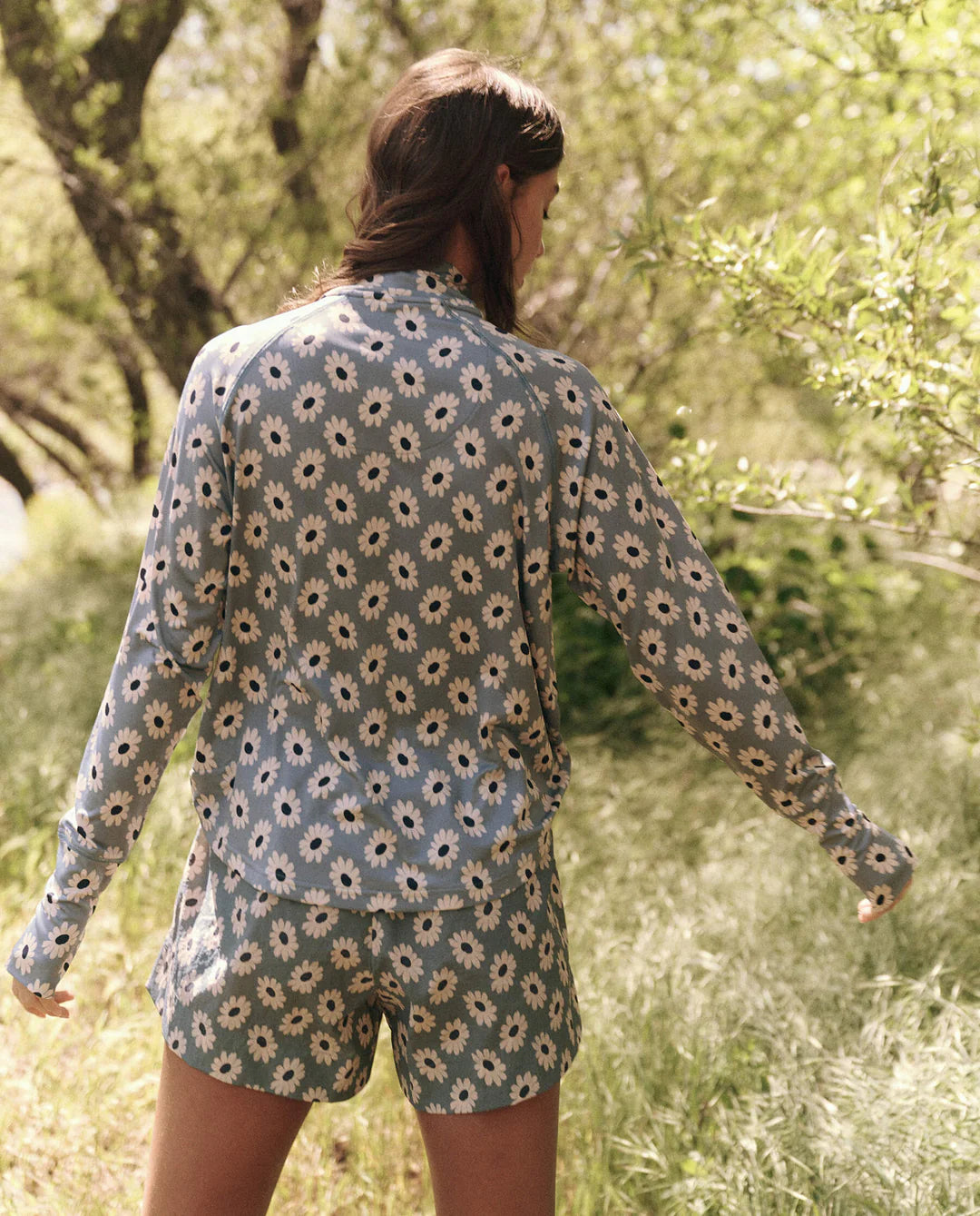 A person outdoors on a grassy field with trees wears The Quarter Zip Trail Pullover from The Great Inc., featuring a blue recycled polyester fabric adorned with white daisy patterns. Sporting a matching floral outfit, the individual faces away from the camera.