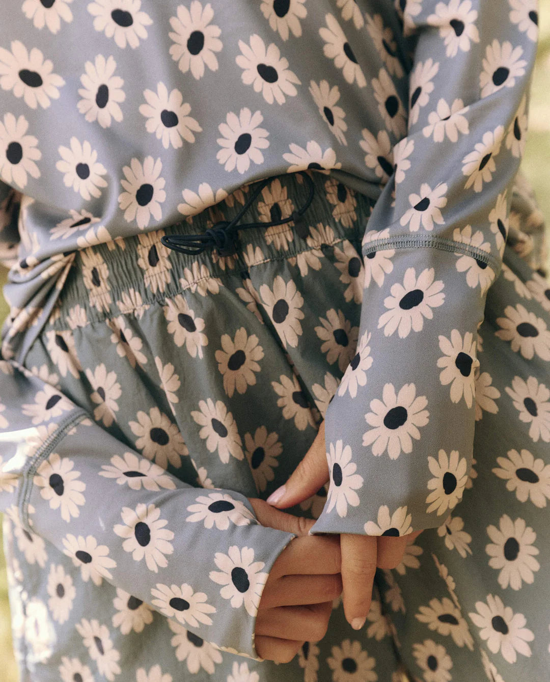 Close-up of a person wearing The Quarter Zip Trail Pullover by The Great Inc. The elegant long-sleeve top, made from recycled polyester, features a muted tone. Hands are gently clasped at the waist, highlighting the eco-conscious design and detailed craftsmanship.